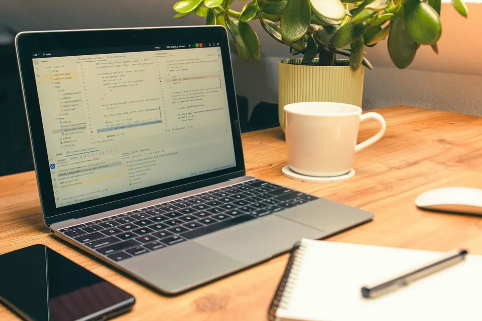 A laptop on a wooden desk with source code on the screen, next to a plant and a cup.